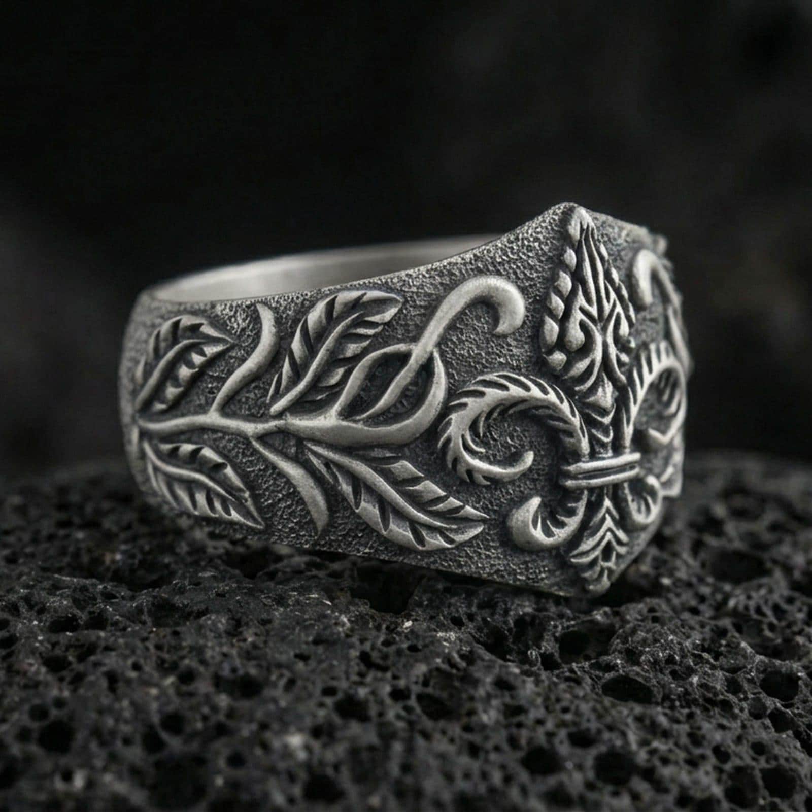 Oxidized sterling silver fleur-de-lis signet ring with engraved vine leaves and textured band, macro photo on black volcanic rock