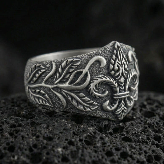 Oxidized sterling silver fleur-de-lis signet ring with engraved vine leaves and textured band, macro photo on black volcanic rock