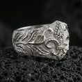 Load image into Gallery viewer, Polished silver fleur-de-lis ring featuring raised leaf scrollwork and a rugged matte texture, close-up product shot on volcanic stone
