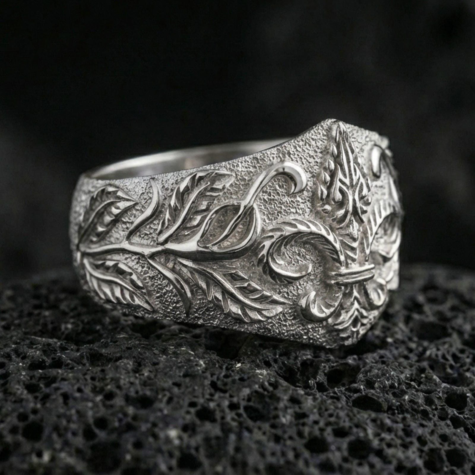 Polished silver fleur-de-lis ring featuring raised leaf scrollwork and a rugged matte texture, close-up product shot on volcanic stone