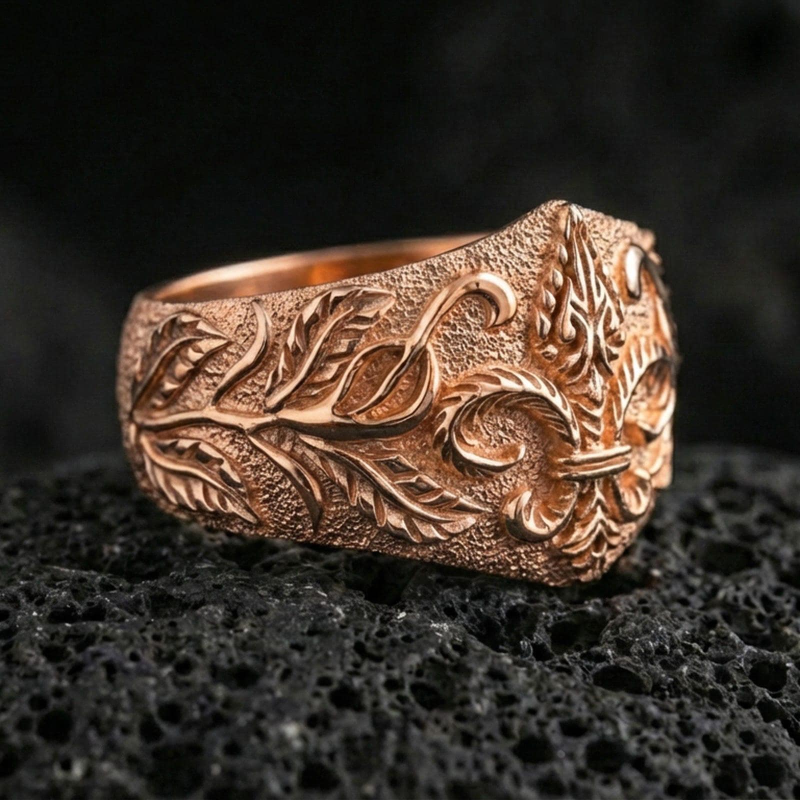 Rose gold plated fleur-de-lis ring with ornate botanical scrolls and vintage-style texture, close-up photo on volcanic rock
