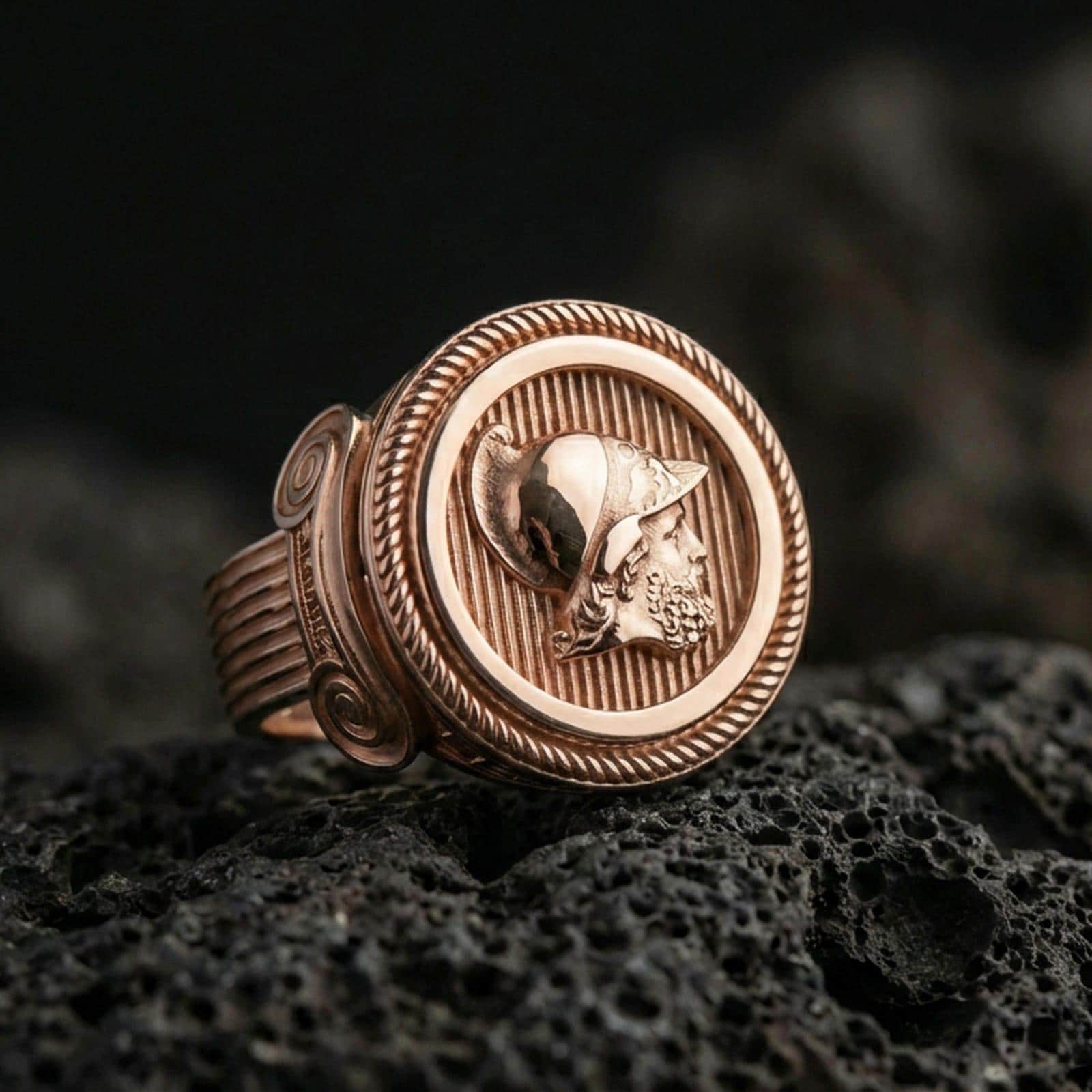 Rose gold plated Greek coin ring with helmeted warrior portrait and rope bezel detail, close-up photo on volcanic rock