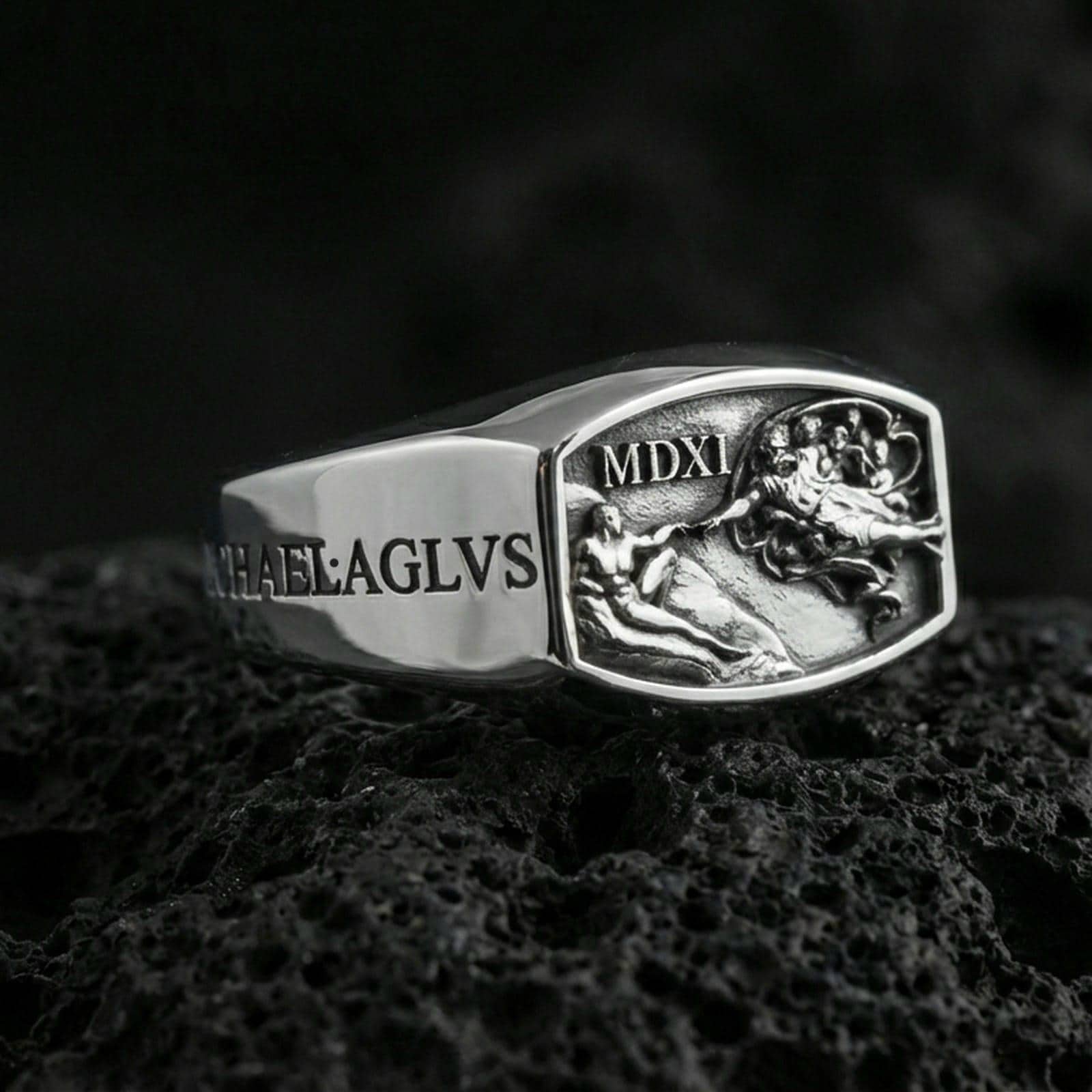 Polished silver Saint Michael ring showing carved “MICHAEL” text and high-relief heavenly battle scene, close-up on volcanic stone