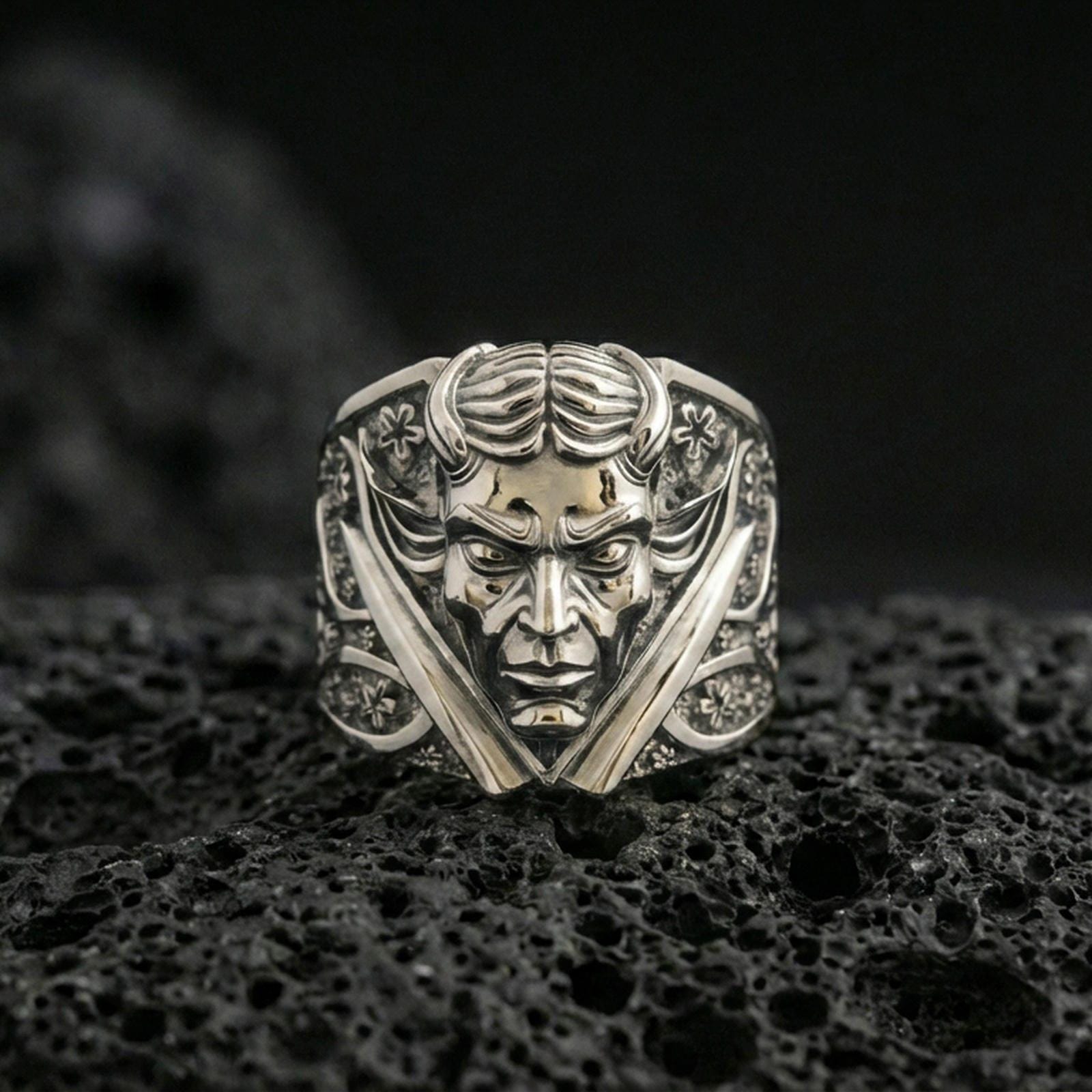 Polished silver horned demon face ring featuring twin daggers and engraved side panels, close-up product shot on volcanic stone