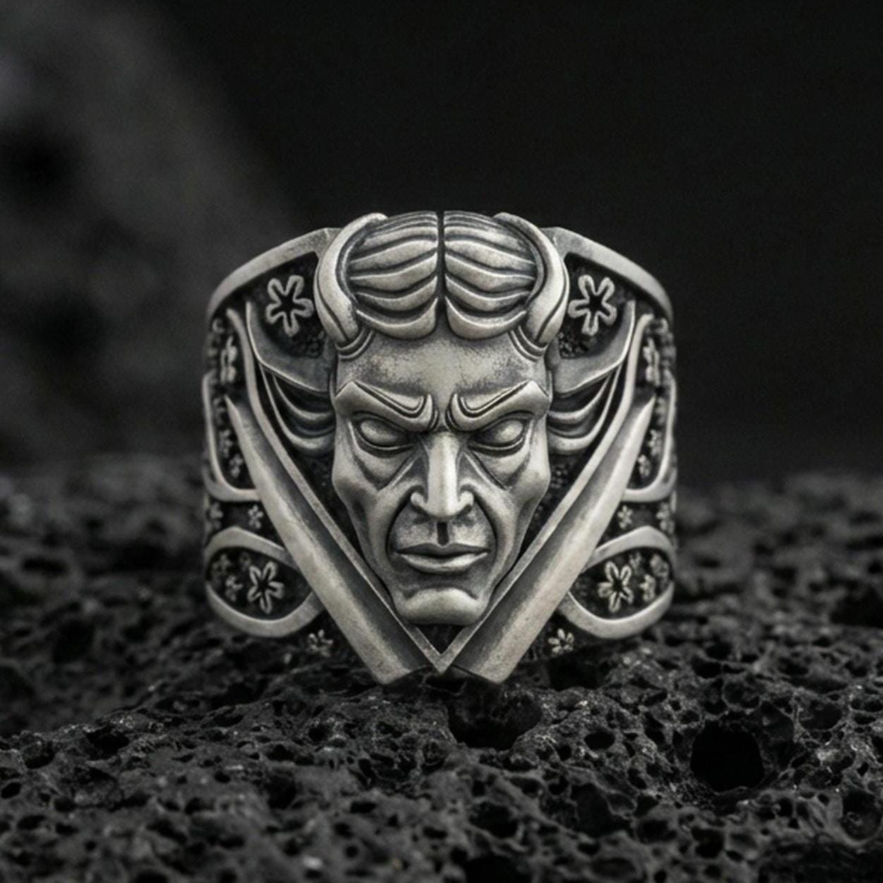 Oxidized sterling silver devil face signet ring with crossed blades and ornate floral accents, macro product photo on black volcanic rock
