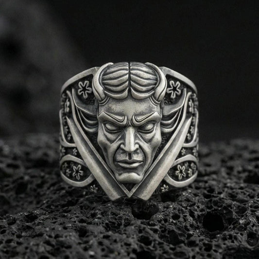 Oxidized sterling silver devil face signet ring with crossed blades and ornate floral accents, macro product photo on black volcanic rock