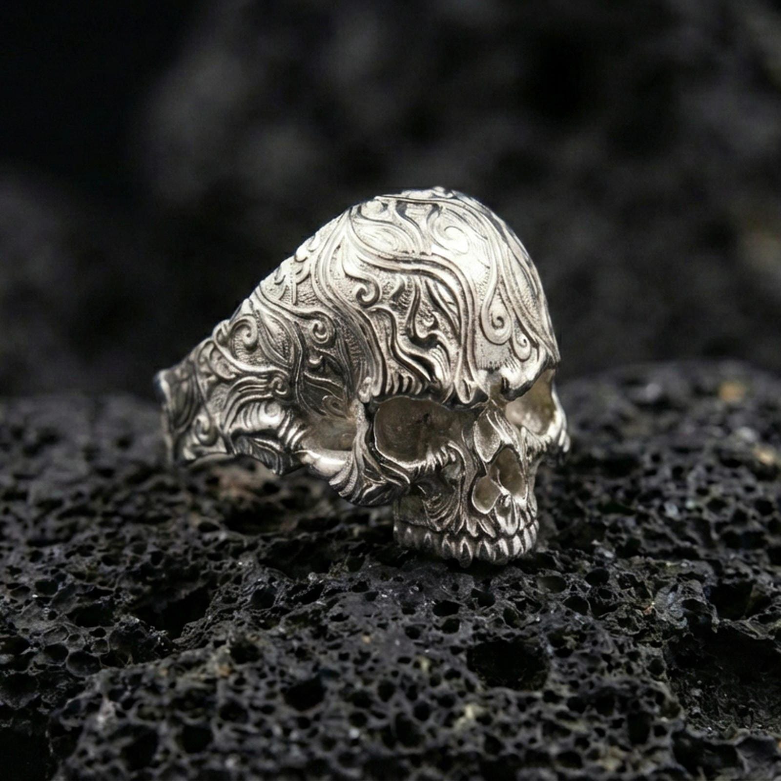 Polished silver gothic skull signet ring featuring ornate floral filigree carving across the band, close-up on volcanic stone