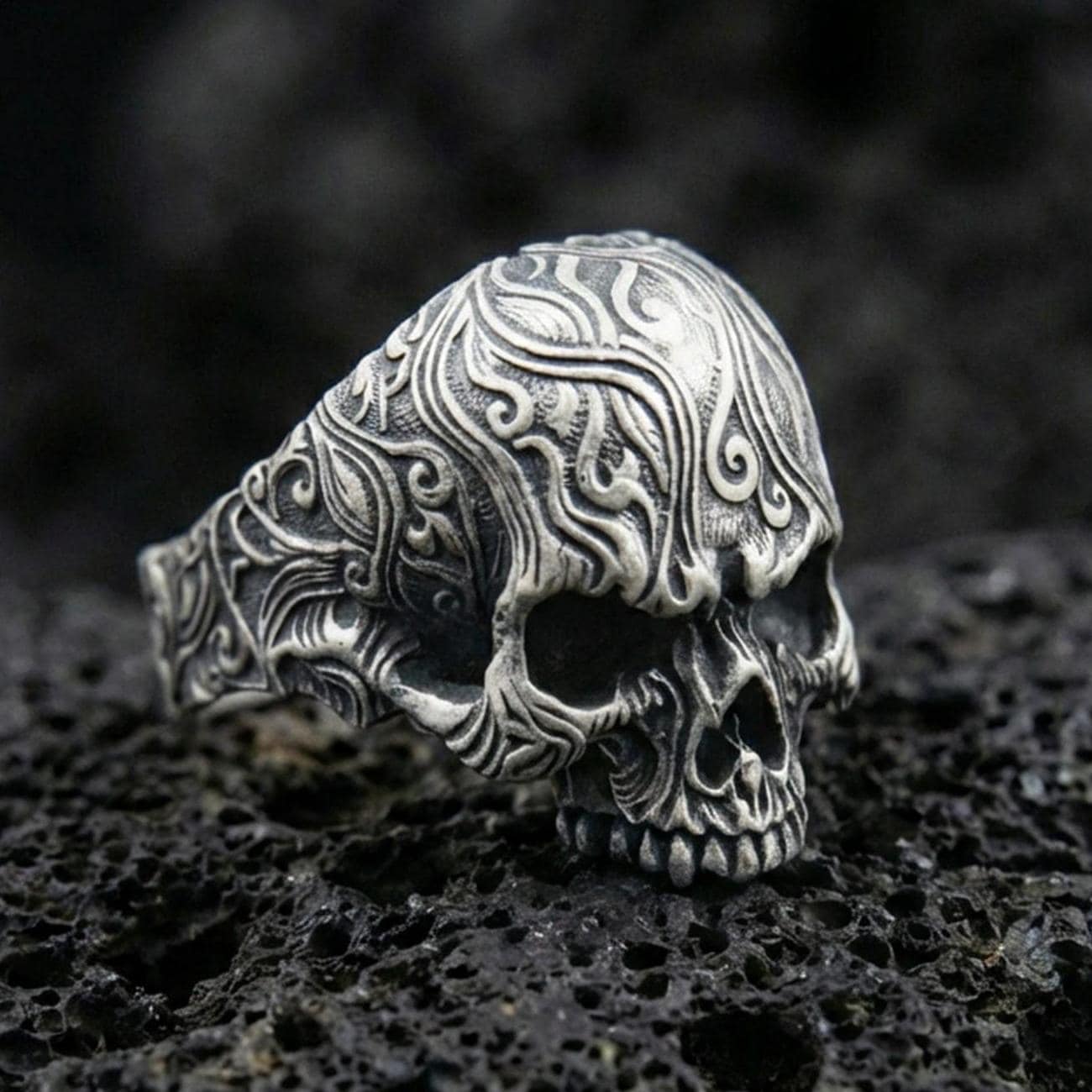 Oxidized sterling silver skull ring with baroque scroll engravings and deep shadowed relief, macro product photo on black volcanic rock