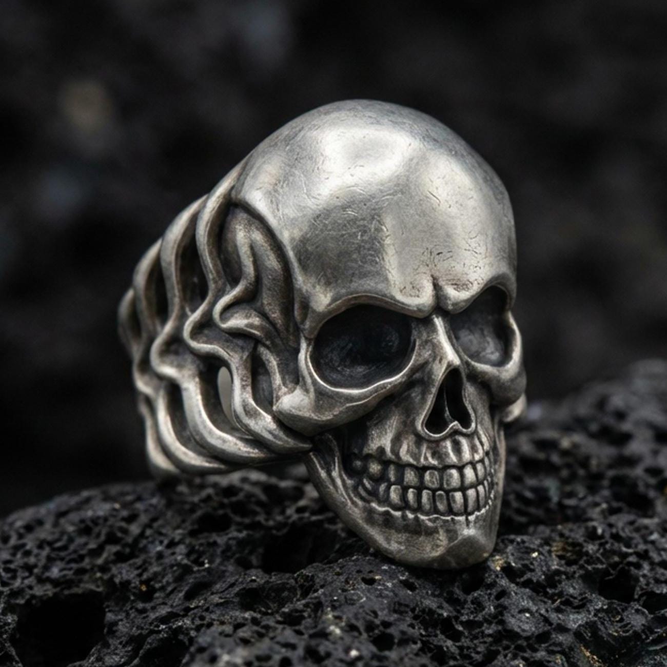 Oxidized sterling silver skull ring with smooth dome forehead and deep blackened eye sockets, macro product photo on black volcanic rock