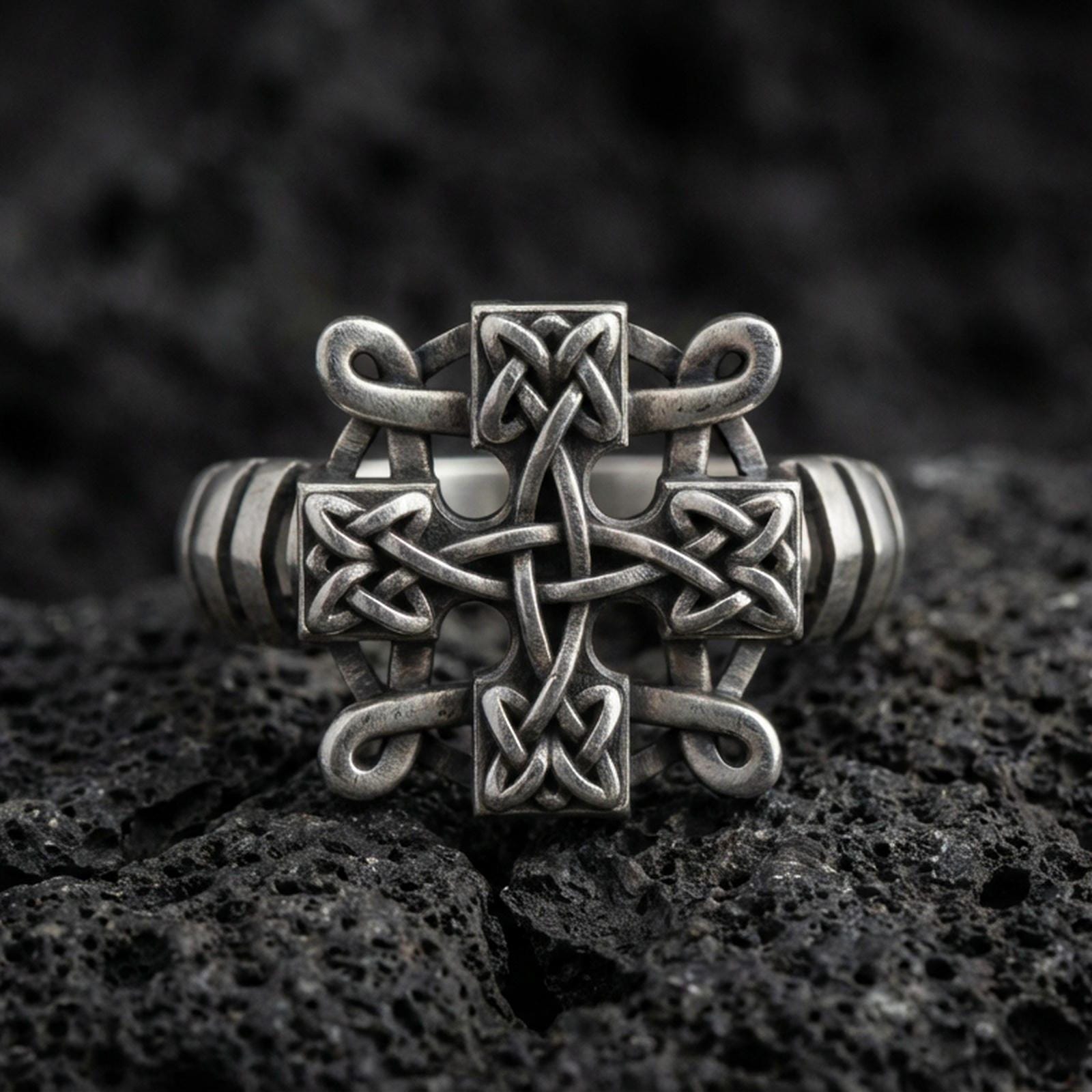 Oxidized sterling silver Celtic knot cross ring with interlaced Trinity-style weave and darkened recessed texture, macro product photo on black volcanic rock
