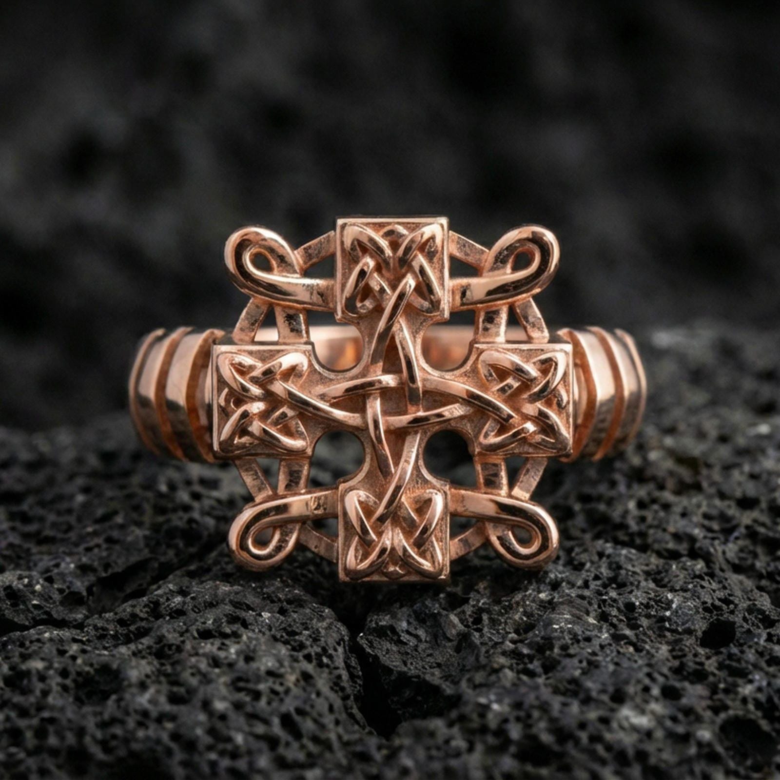 Rose gold plated Celtic knot cross ring with intricate interlace pattern and sculpted cross silhouette, close-up on volcanic rock