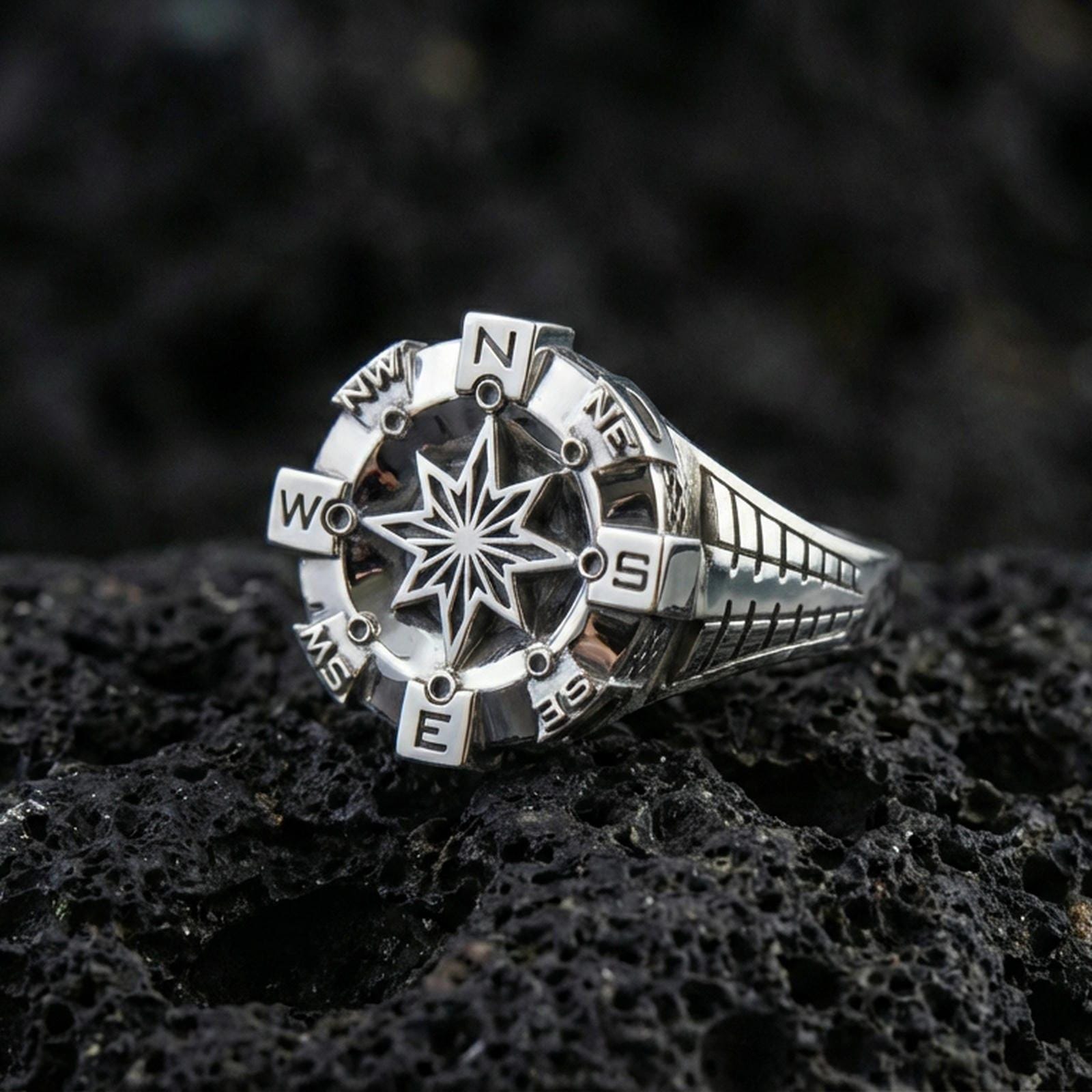 Polished silver compass signet ring featuring a dimensional navigation star and engraved direction letters, close-up on volcanic stone