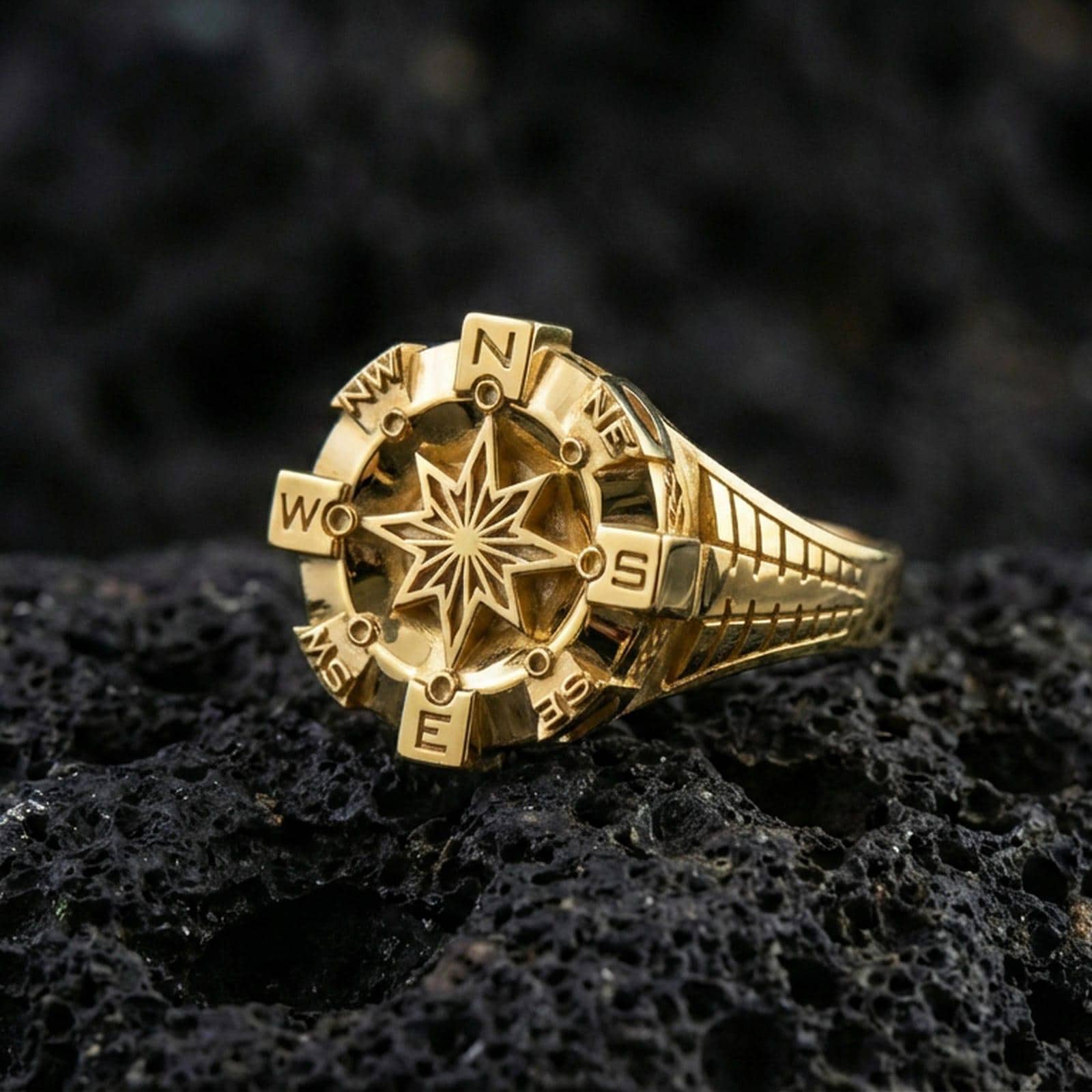 Gold plated compass ring with bold eight-point star centerpiece and directional initials around the bezel, product-only macro image on black volcanic rock