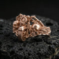 Load image into Gallery viewer, Rose gold plated biker skull ring with dual skull faces, handlebar centerpiece, and rugged chain accents, close-up product shot on volcanic rock
