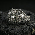 Load image into Gallery viewer, Polished silver biker ring featuring two skulls, a motorcycle handlebar centerpiece, and heavy chain detailing, close-up on volcanic stone
