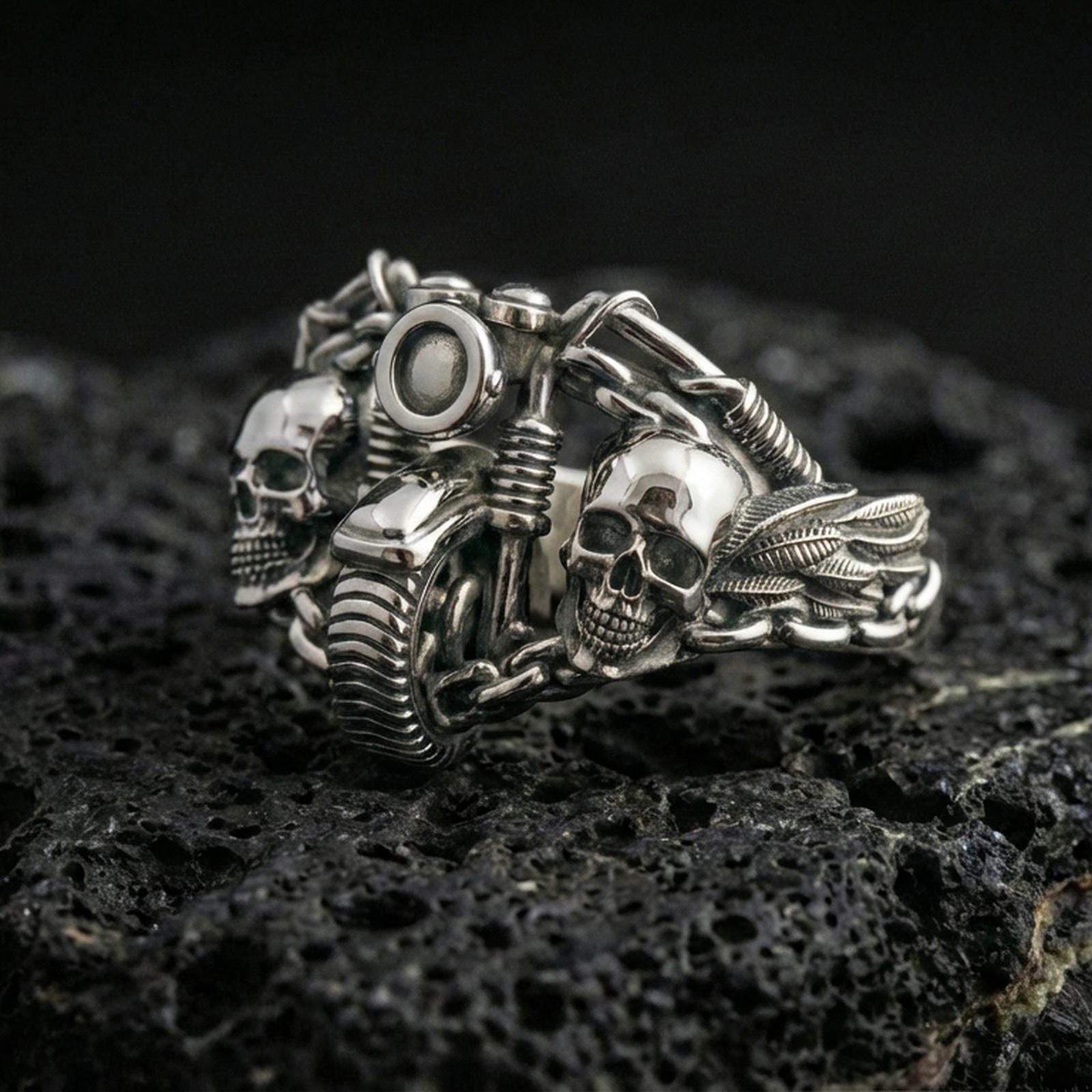 Polished silver biker ring featuring two skulls, a motorcycle handlebar centerpiece, and heavy chain detailing, close-up on volcanic stone