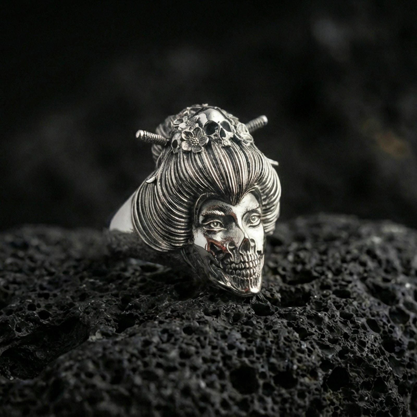 Polished silver Japanese geisha skull ring featuring sculpted facial detail, ribbed hair texture, and hairpin accents, close-up on volcanic stone