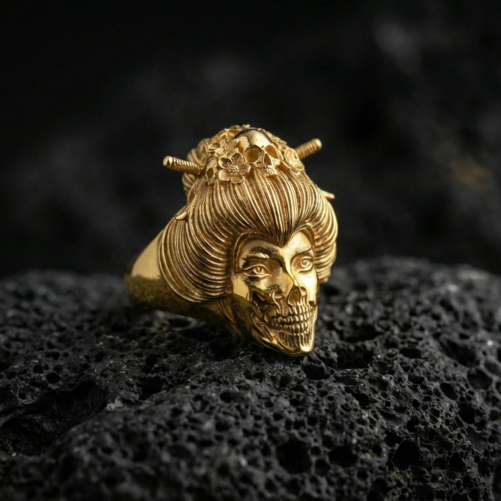 Gold plated geisha skull ring with engraved hair strands, floral crown details, and skull face design, product-only macro image on black volcanic rock
