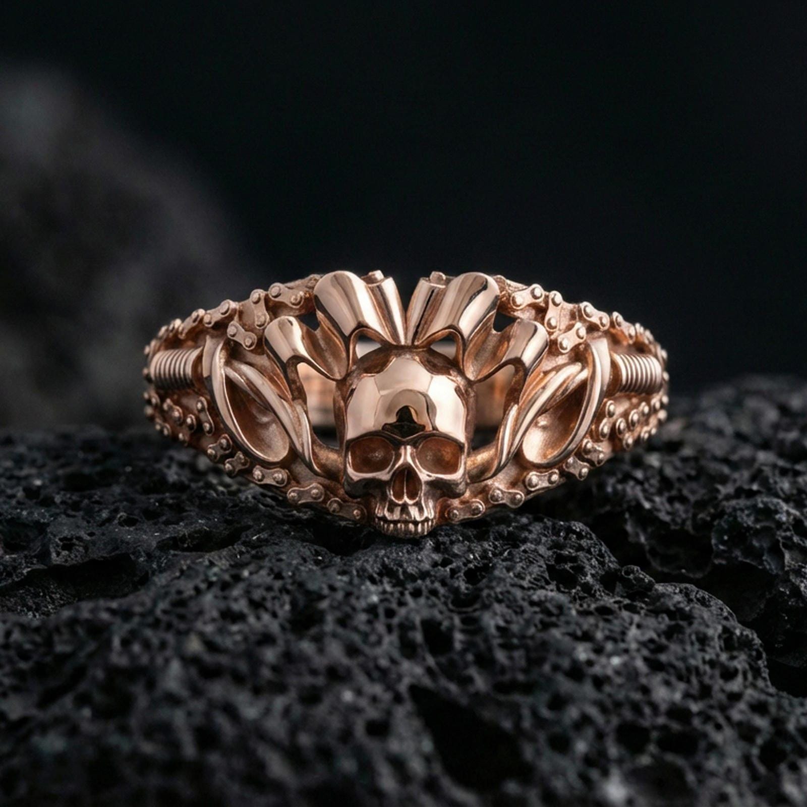 Rose gold plated skull ring with crowned skull centerpiece and detailed chain-link sides, close-up product shot on black volcanic rock