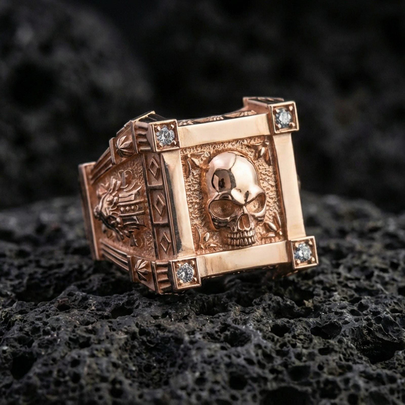 Rose gold plated skull ring with square bezel-style frame, raised skull centerpiece, and lab diamond corners, close-up on black volcanic rock