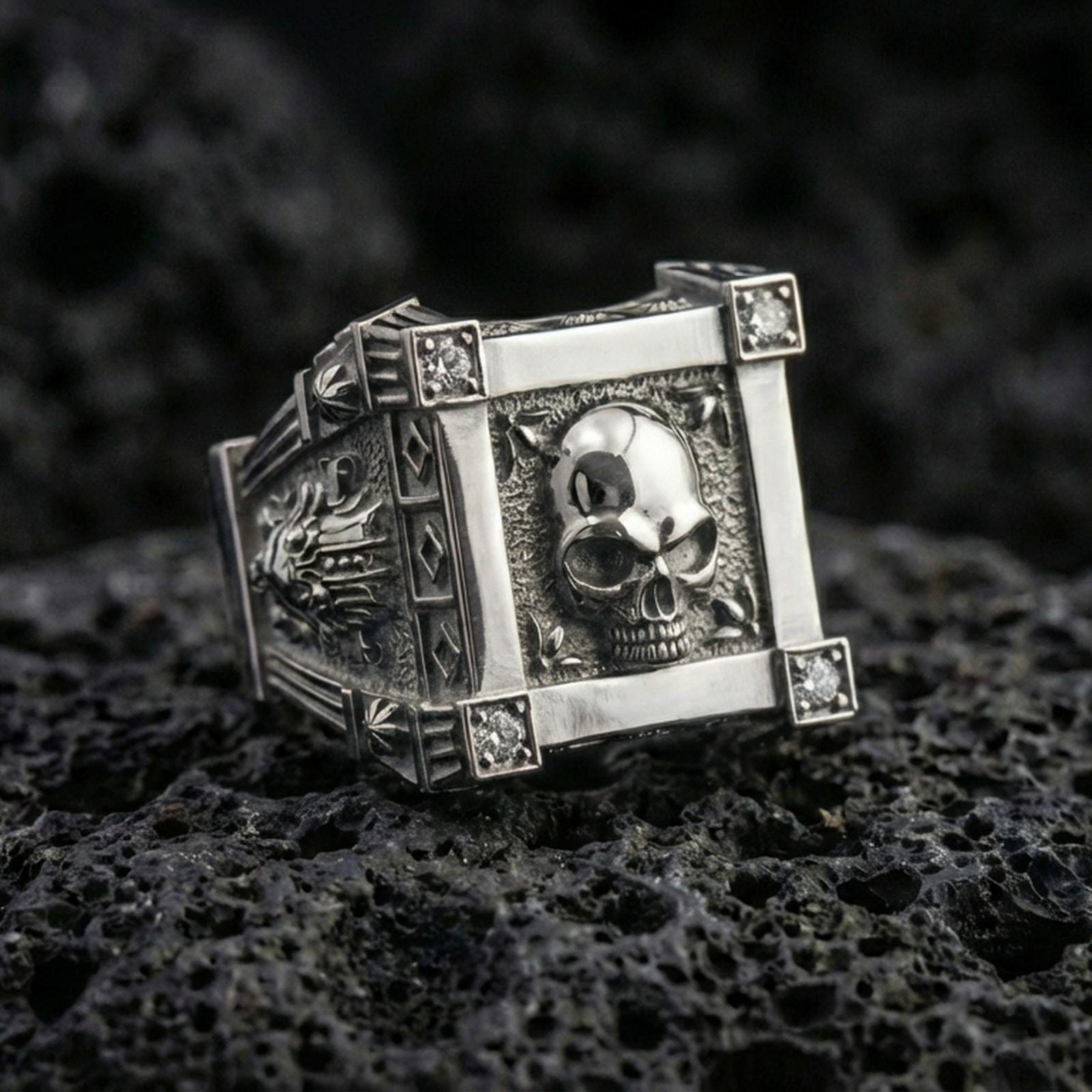 Polished silver skull ring featuring a framed skull centerpiece and lab diamond corner details, close-up on volcanic stone