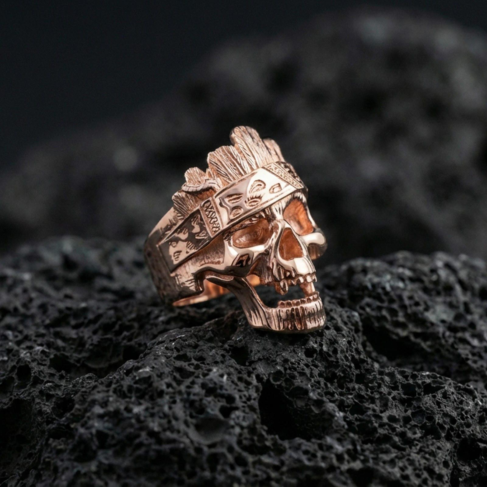 Rose gold plated skull ring with tree bark texture carving and engraved band motifs, close-up on black volcanic rock