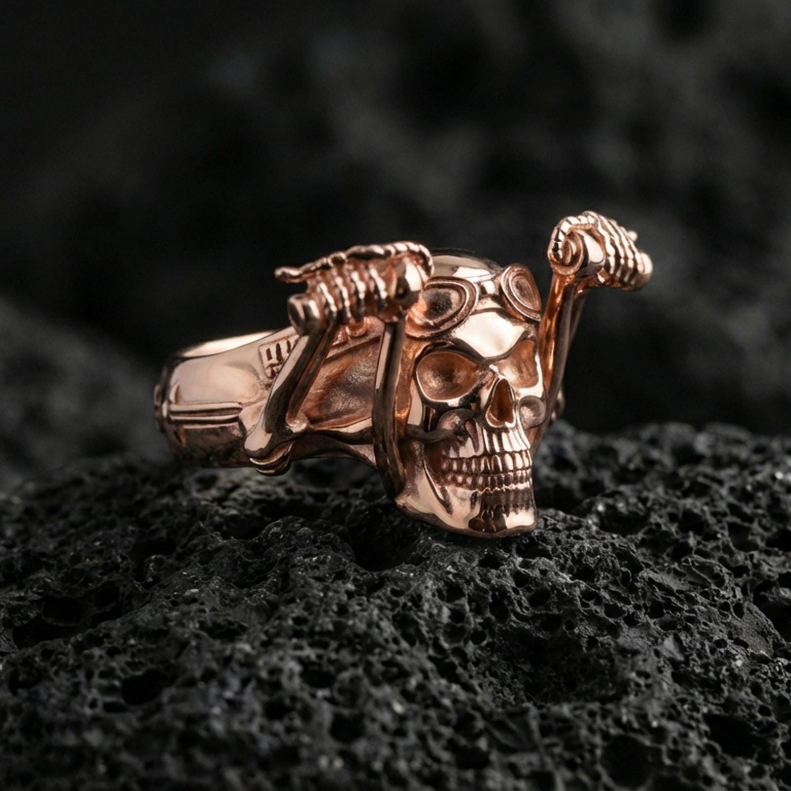 Rose gold plated skull biker ring with aviator goggles and raised skeleton arms, close-up on black volcanic rock