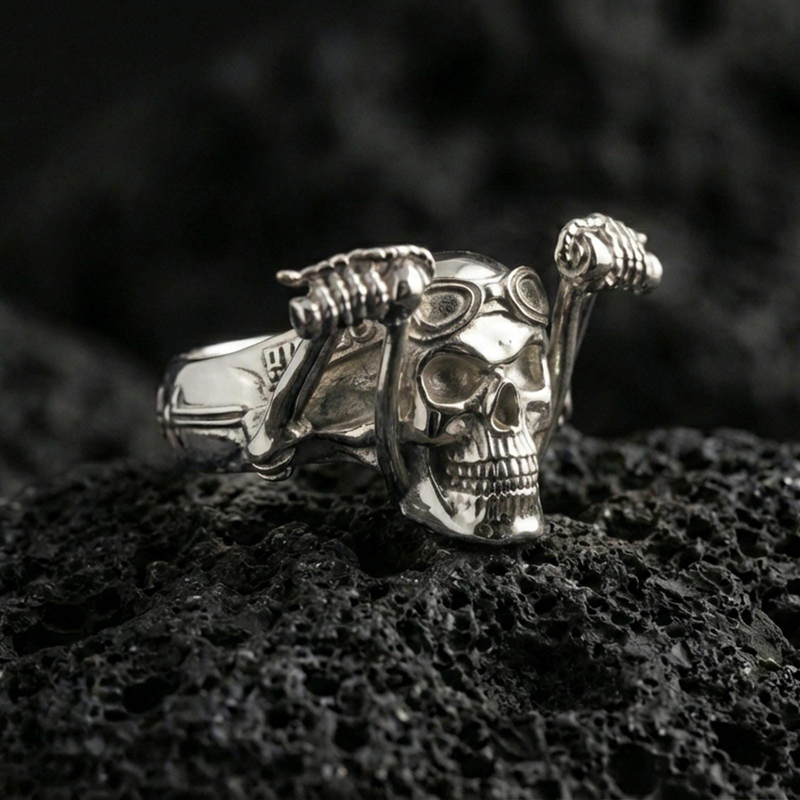 Polished silver skull biker ring featuring aviator goggles and sculpted skeleton arms, close-up on volcanic stone