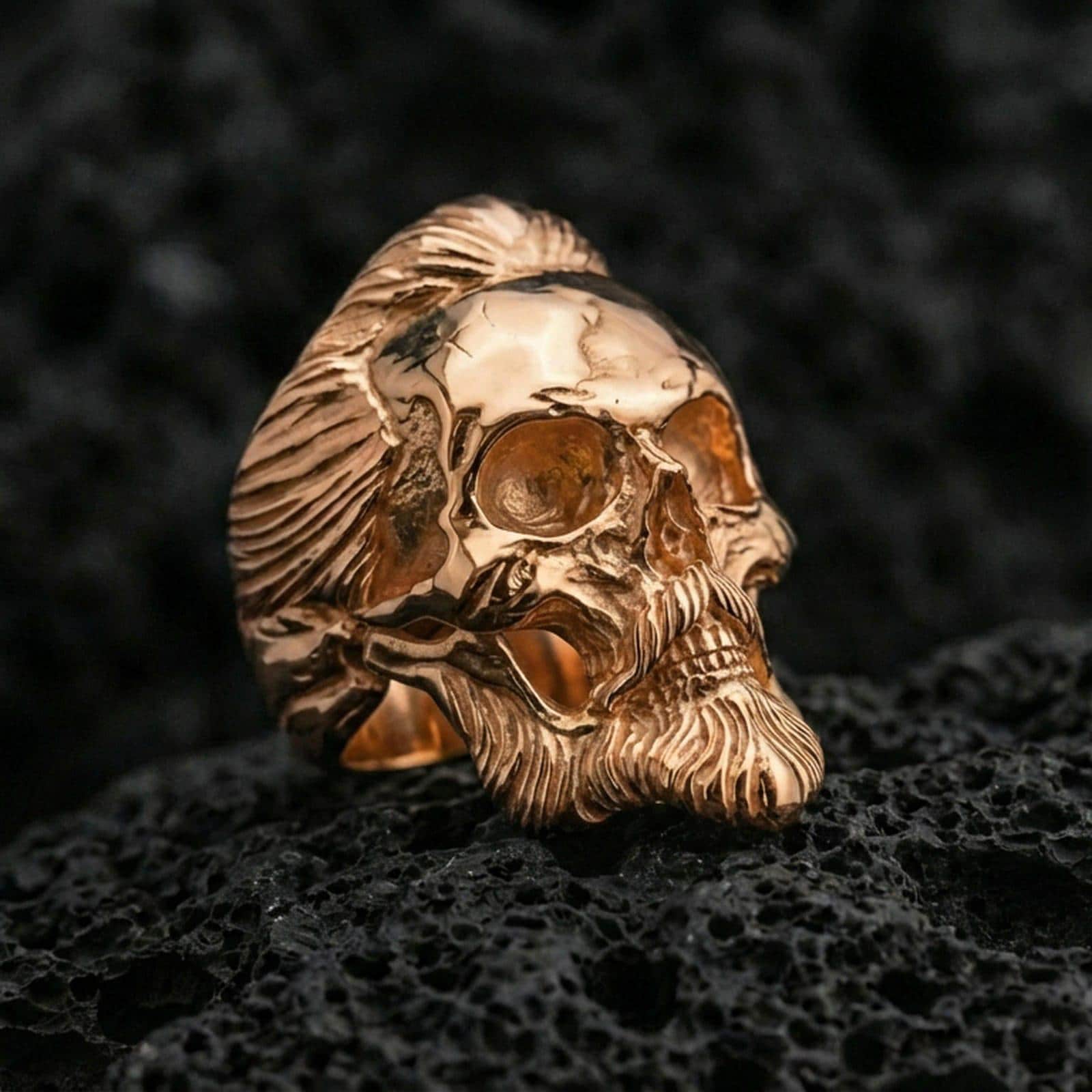 Rose gold plated bearded skull ring with high-relief hair and beard detailing, close-up on volcanic rock