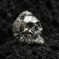 Load image into Gallery viewer, Polished silver bearded skull ring with detailed hair texture and deep eye sockets, close-up on volcanic stone
