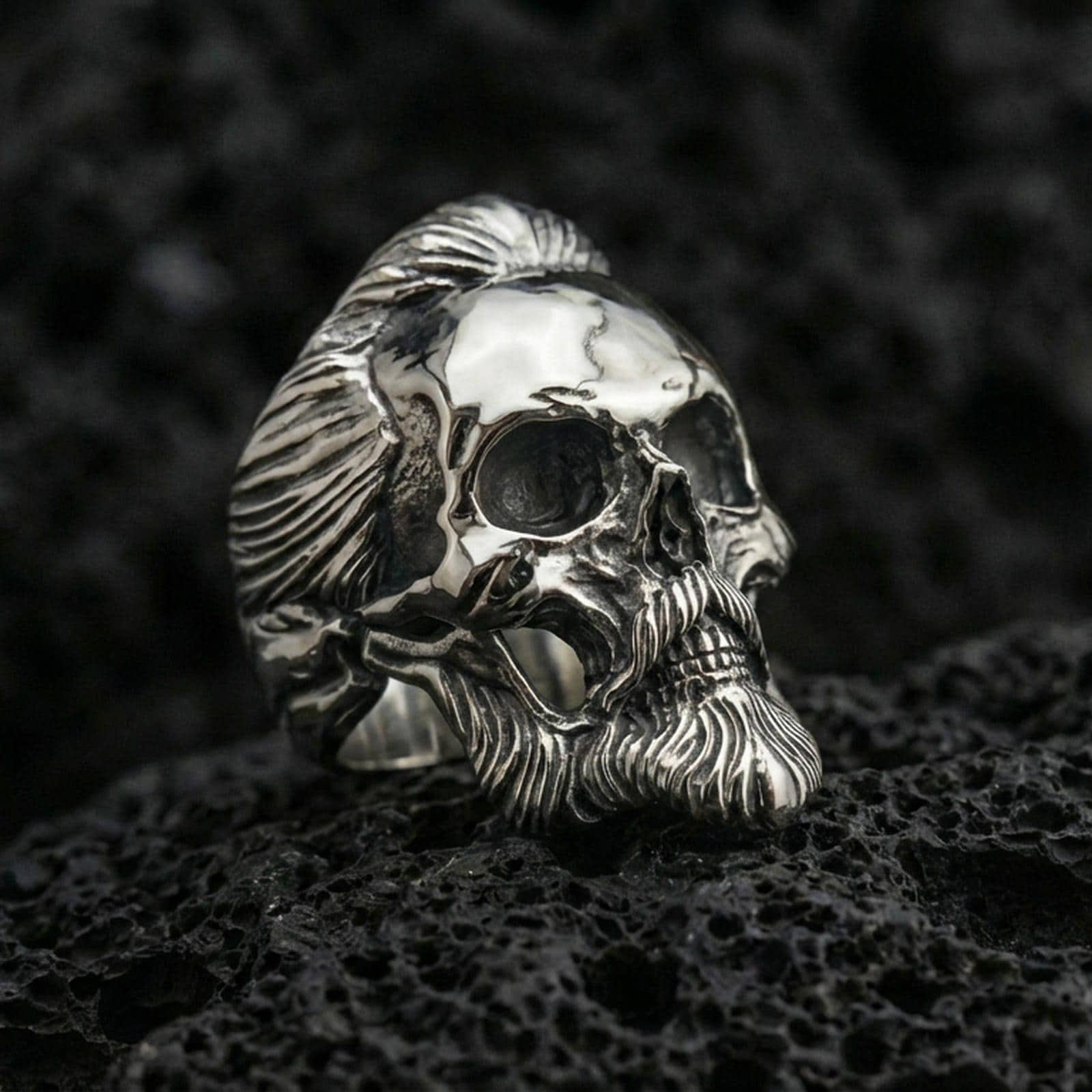 Polished silver bearded skull ring with detailed hair texture and deep eye sockets, close-up on volcanic stone