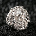 Load image into Gallery viewer, Polished silver ornate skull ring featuring flowing smoke scrolls and spiral eye detail, close-up on volcanic stone
