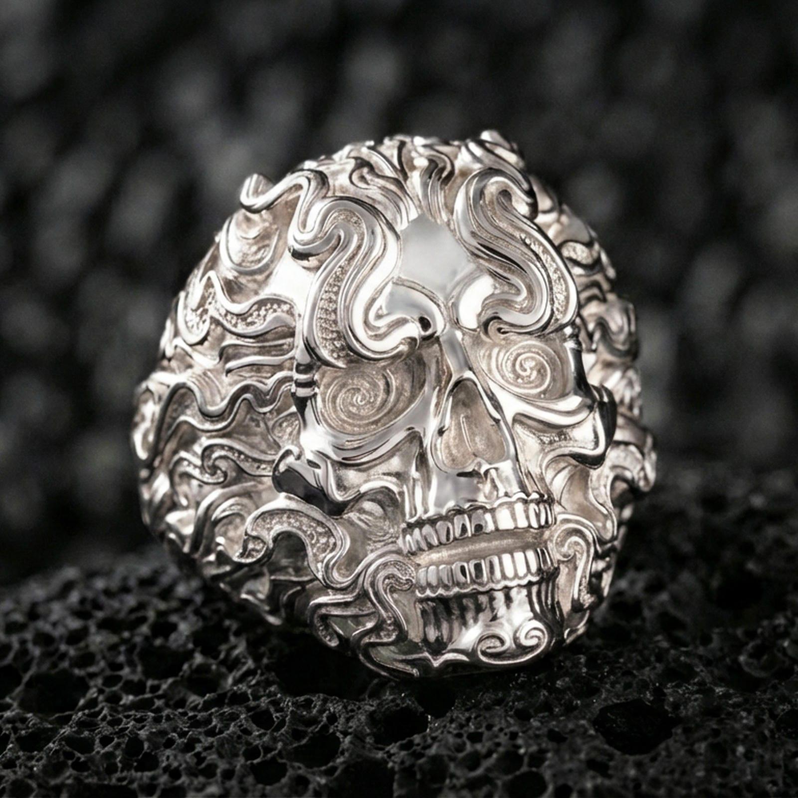Polished silver ornate skull ring featuring flowing smoke scrolls and spiral eye detail, close-up on volcanic stone