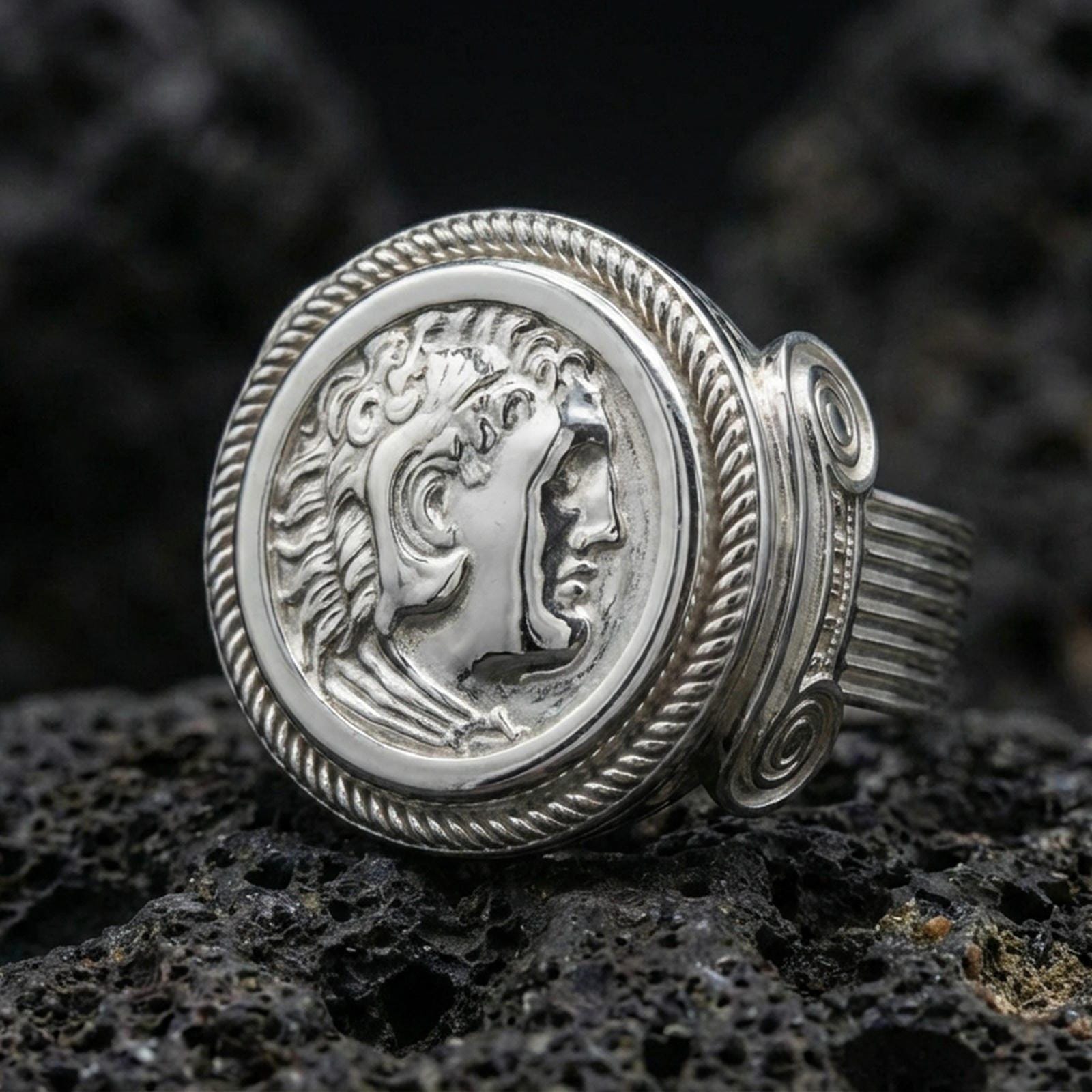 Polished silver ancient coin signet ring with a Roman-style emperor profile and twisted rope bezel, close-up on volcanic stone