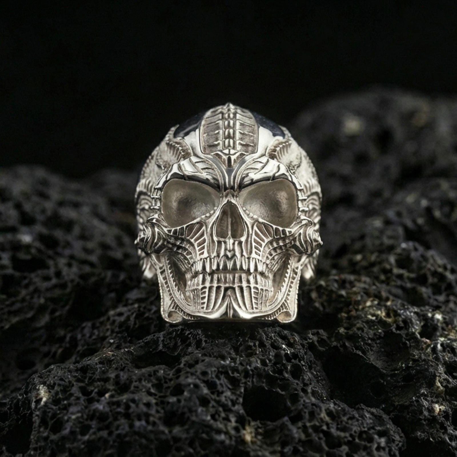 Polished silver cyber skull ring featuring mechanical detailing, hollow eye sockets, and sharp contour lines, close-up on volcanic stone