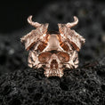 Load image into Gallery viewer, Rose gold plated horned skull ring with dramatic curved horns and bold skull centerpiece, close-up on volcanic rock
