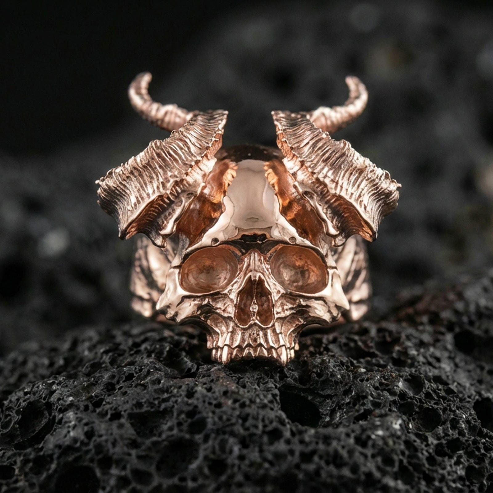 Rose gold plated horned skull ring with dramatic curved horns and bold skull centerpiece, close-up on volcanic rock