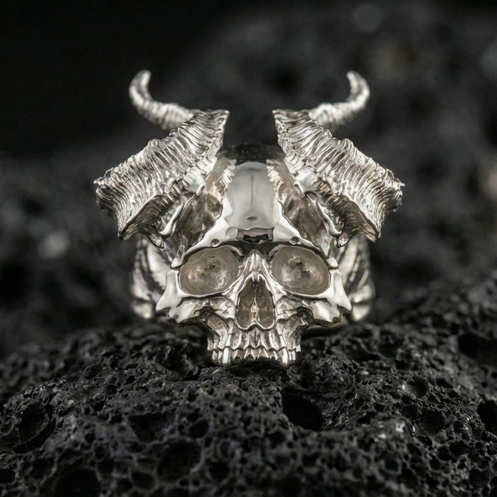 Polished silver horned skull statement ring with textured curved horns and sculpted skull face, close-up on volcanic stone