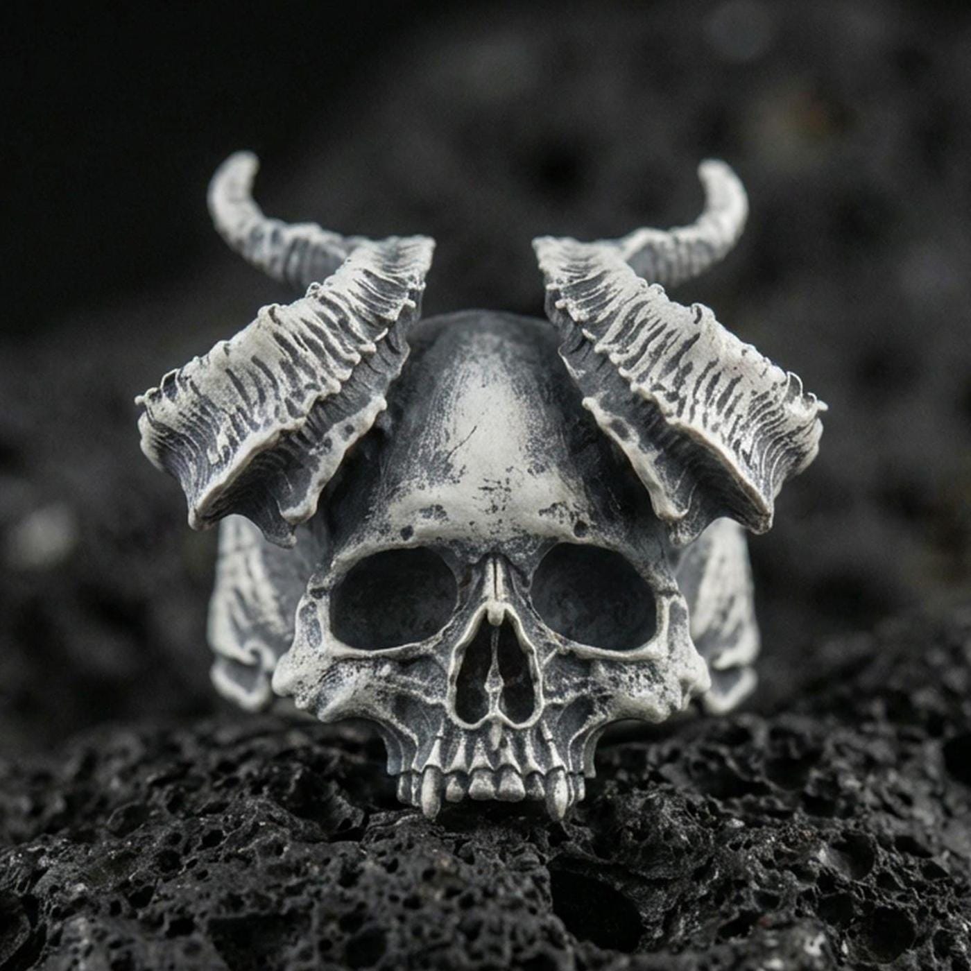 Oxidized sterling silver horned skull ring with curved ram-style horns and deep shadowed details, macro photo on black volcanic rock