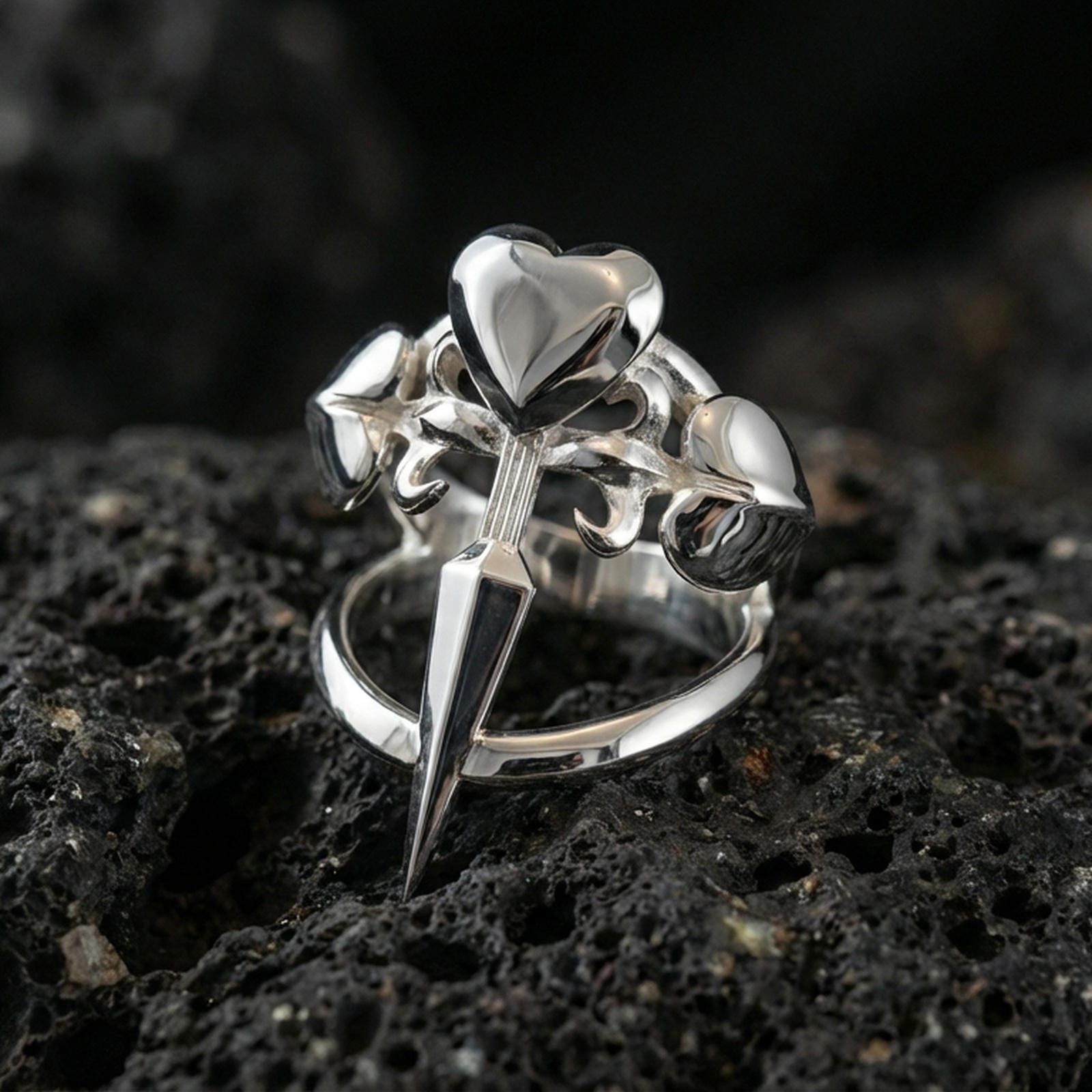 Polished silver heart dagger ring featuring a sculpted blade and three heart motifs, close-up product shot on volcanic stone