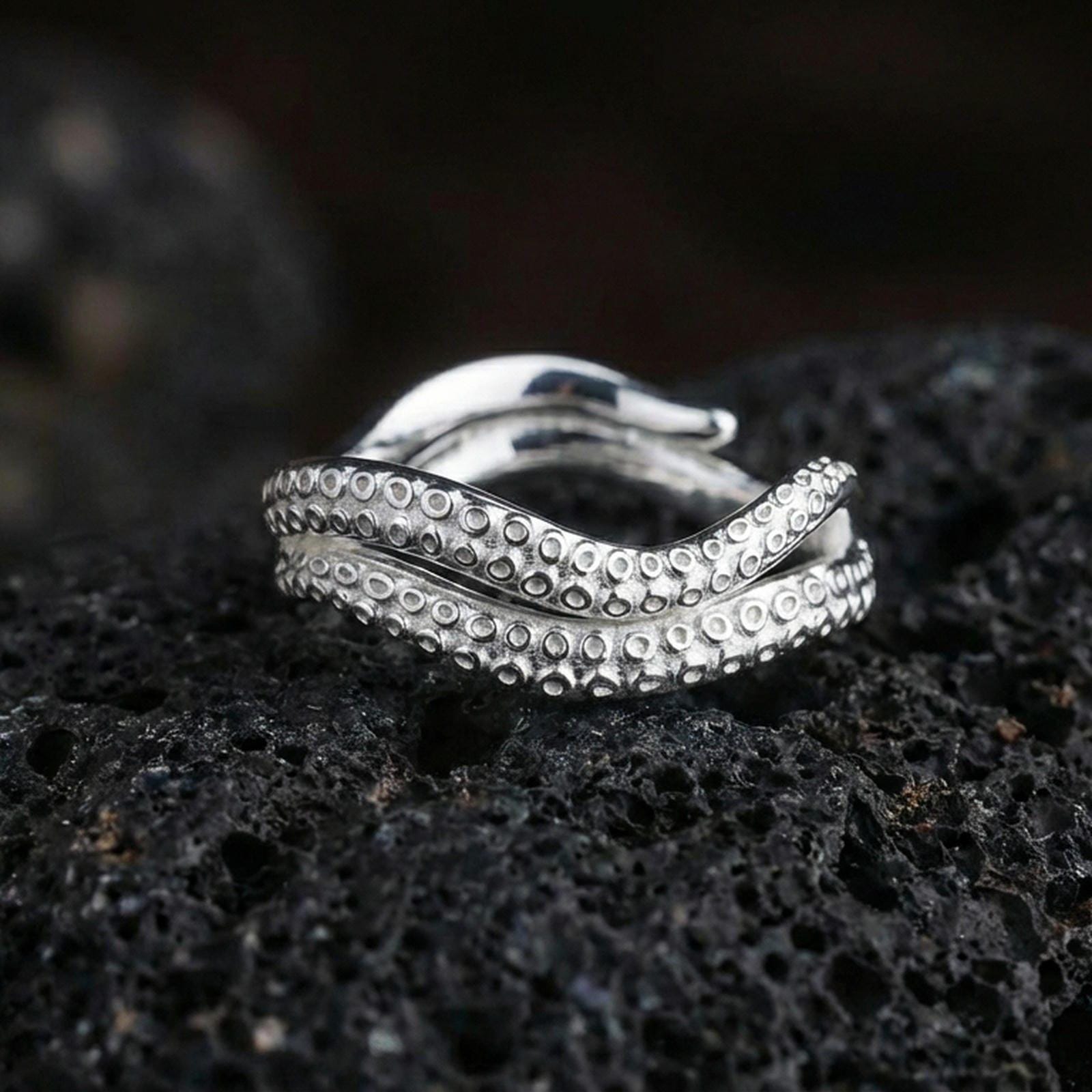 Polished silver octopus tentacle open band ring on black volcanic stone, crisp suction cup engraving and curved wrap design, centered close-up studio lighting.