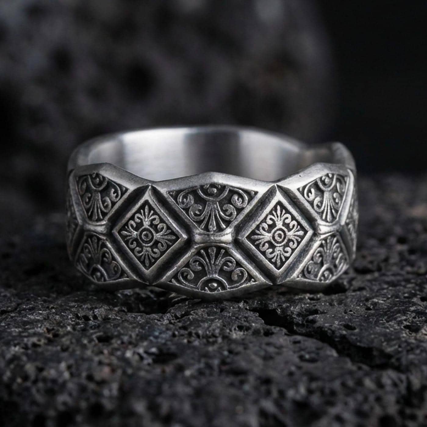 Oxidized sterling silver engraved ornament band ring on dark volcanic rock, raised diamond panels with scrollwork detail, premium macro product shot.