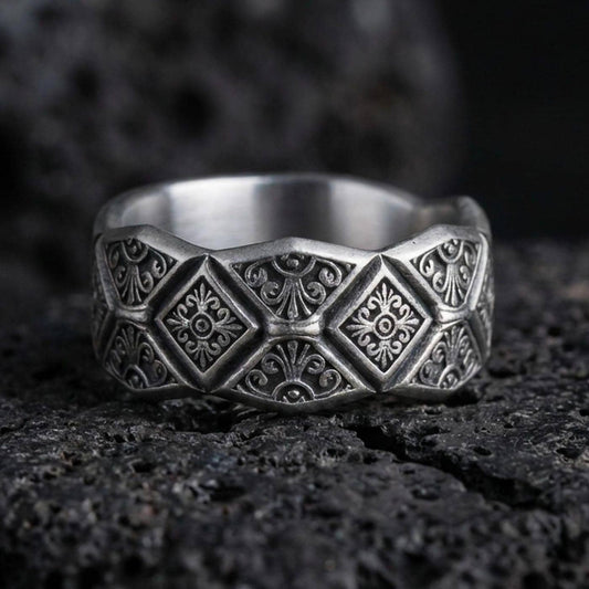 Oxidized sterling silver engraved ornament band ring on dark volcanic rock, raised diamond panels with scrollwork detail, premium macro product shot.