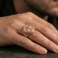 Load image into Gallery viewer, Rose gold plated floral signet ring on the finger, close-up lifestyle image focusing on the rosette medallion and patterned shoulders
