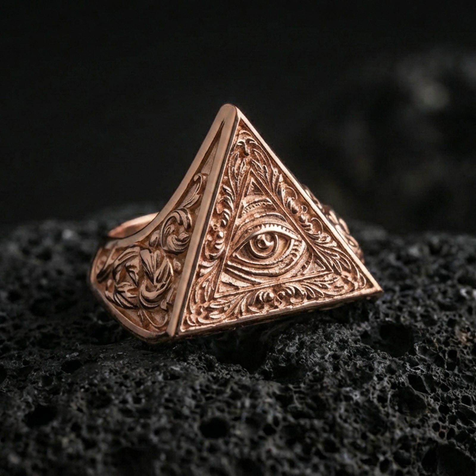 Rose gold plated pyramid triangle signet ring with all-seeing eye emblem and ornamental scrollwork, close-up on volcanic rock