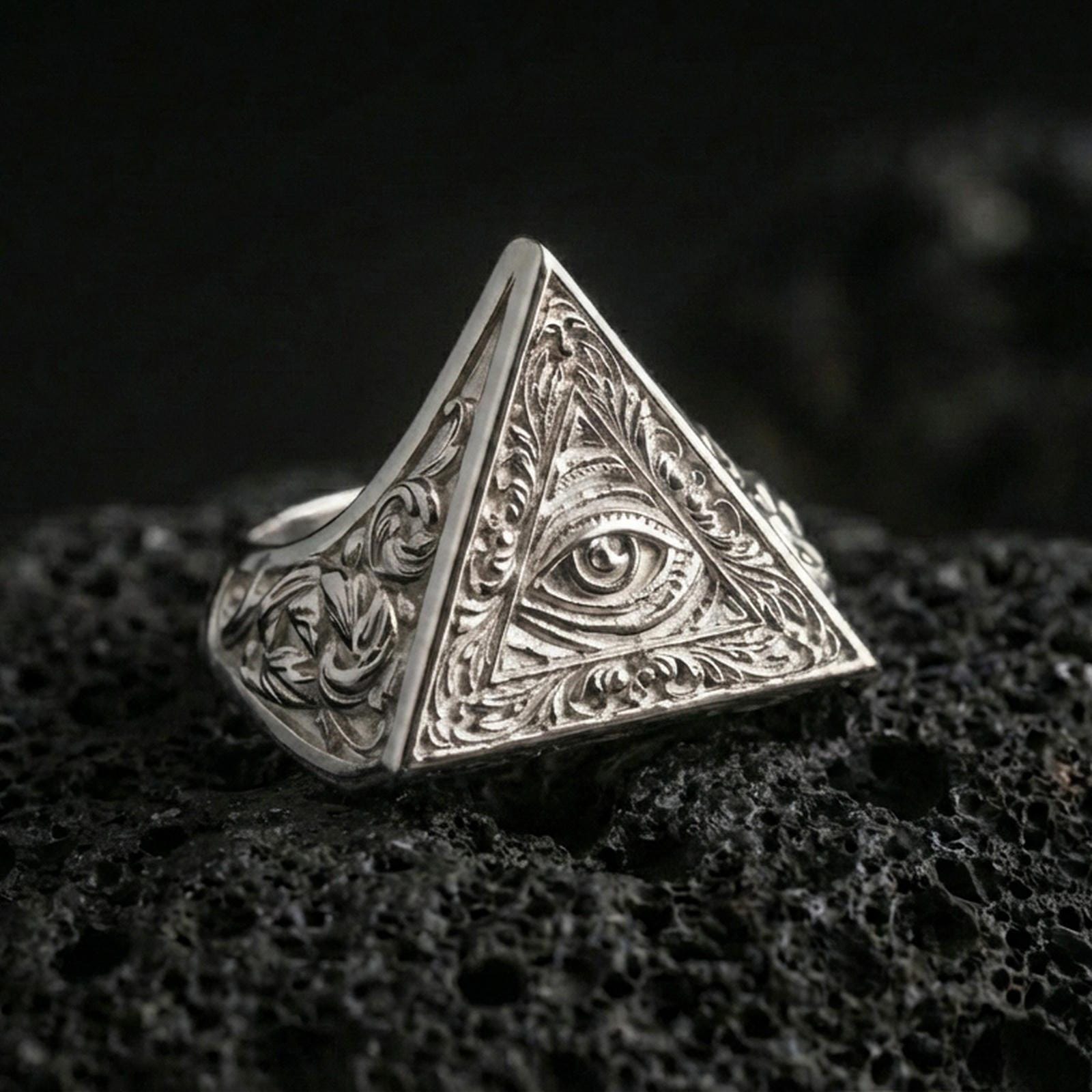 Polished silver pyramid signet ring featuring an engraved all-seeing eye inside a triangle, close-up on volcanic stone