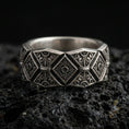 Load image into Gallery viewer, Oxidized sterling silver engraved ornament band ring on dark volcanic rock, bold raised diamond panels with filigree scrollwork, premium macro product shot.
