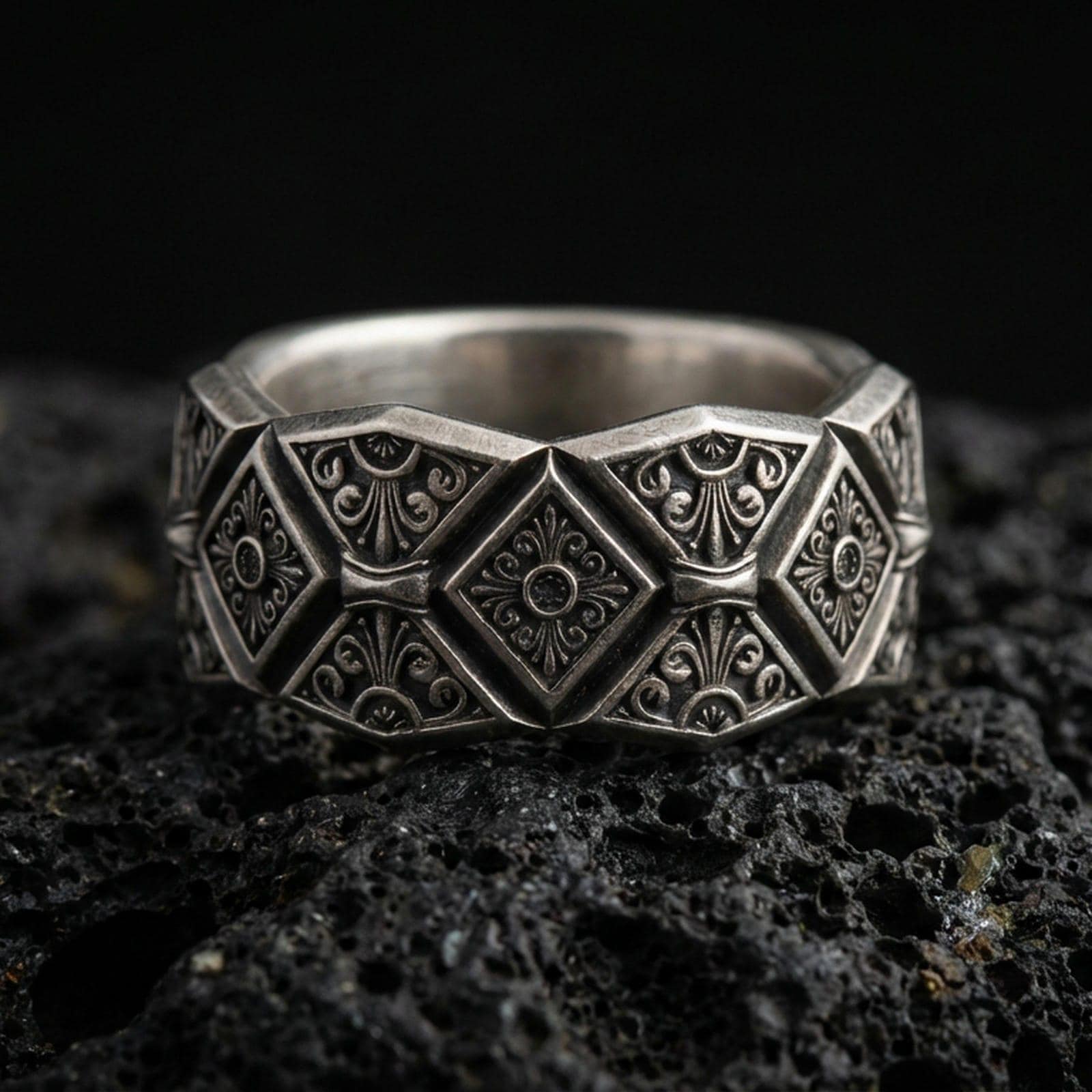 Oxidized sterling silver engraved ornament band ring on dark volcanic rock, bold raised diamond panels with filigree scrollwork, premium macro product shot.