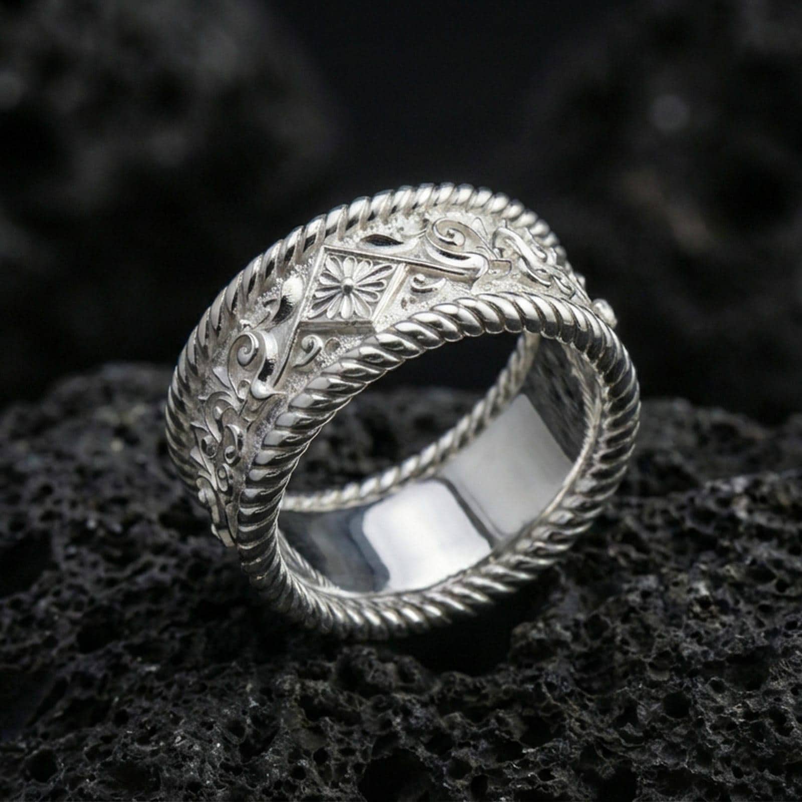 Polished silver rope border engraved band ring on black volcanic stone, crisp floral panel and vintage scrollwork detail, centered close-up studio lighting.