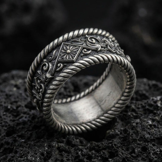 Oxidized sterling silver rope edge band ring on dark volcanic rock, ornate floral and scroll engraving, premium macro product shot.